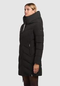 Zwarte pufferjas met een hoge kraag, chevronpatroon, ritszakken en logotag. Heeft een getailleerde pasvorm en textuurstof.