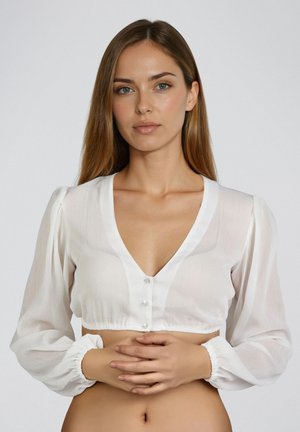 Frau mit hellbraunen Haaren trägt eine weiße, gekürzte Bluse mit Knöpfen, steht mit verschränkten Händen auf Taillenhöhe vor einem schlichten Hintergrund.