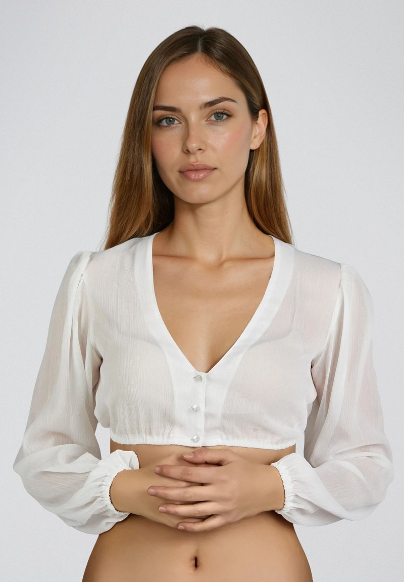 Femme aux cheveux châtain clair portant un chemisier blanc court à boutons, debout les mains jointes à la taille devant un fond uni.