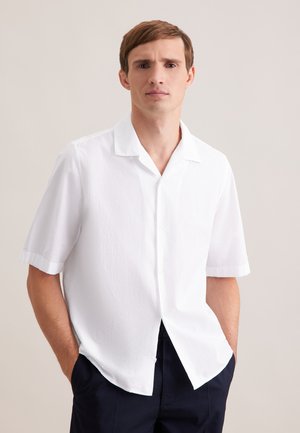 Ung mand med kort brunt hår iført en tekstureret hvid kortærmet skjorte med knapper og mørke bukser, stående med hænderne i lommerne.