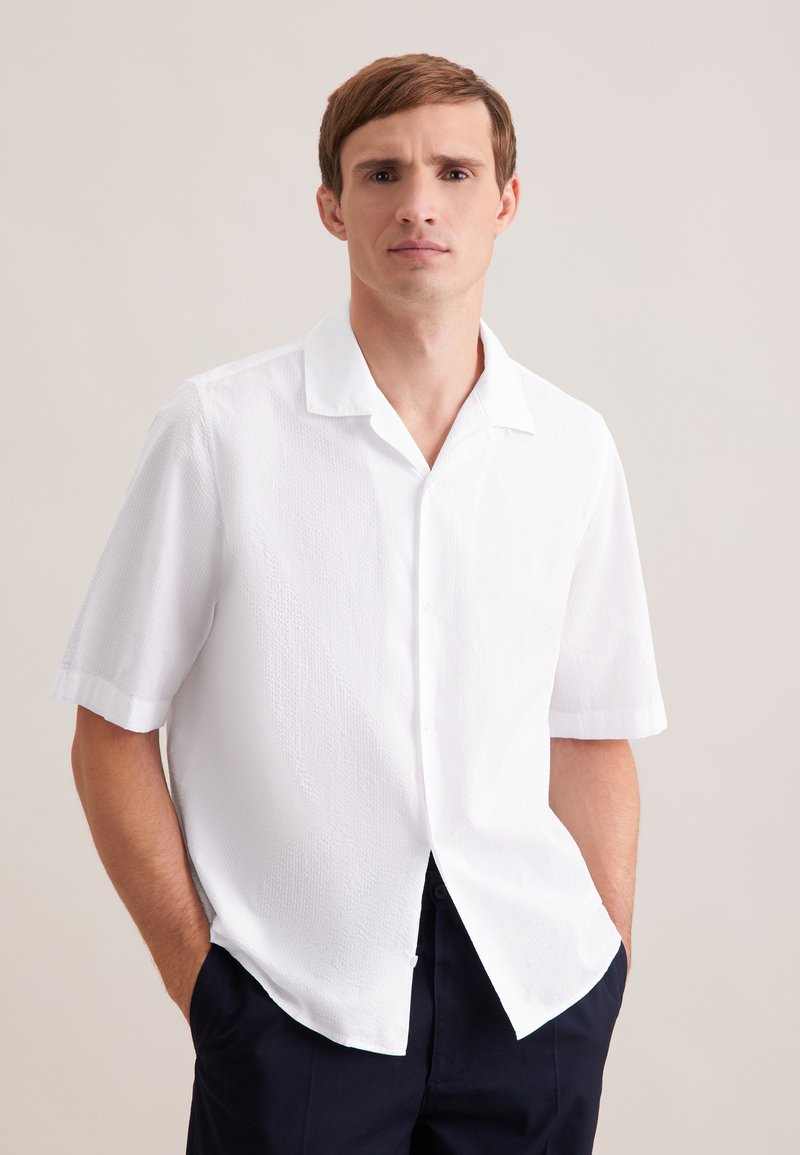 Giovane uomo con capelli castani corti, indossa una camicia bianca a maniche corte con trama e pantaloni scuri, in piedi con le mani nelle tasche.