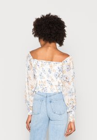 Blusa floreale off-the-shoulder con maniche lunghe a sbuffo, caratterizzata da motivi floreali blu e oro su uno sfondo chiaro. Design corto.