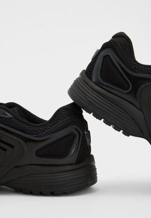 Paar schwarze Sportschuhe mit Mesh- und synthetischen Überzügen, dicke gepolsterte Sohlen, vor einem einfarbigen hellgrauen Hintergrund positioniert.