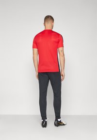 Rotes kurzärmeliges Sportshirt und schwarze tapered Hosen. Das Shirt hat eine glatte Textur; die Hose hat einen schmalen Schnitt mit Knöchelbündchen. Fußballschuhe sind sichtbar.