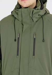 Grüne wasserdichte Jacke mit hohem Kragen, verstellbarem Kapuzen und schwarzen Reißverschlusstaschen. Verfügt über einen glatten, strukturierten Stoff und Kordeln.