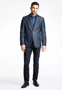 Dunkelblauer Denim-Blazer mit Reverskragen, Ein-Knopf-Verschluss und zwei vorderen Taschen, kombiniert mit dunklen Jeans und schwarzen Schuhen.