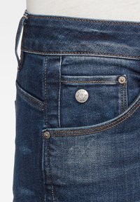 Blue denim jeans with a mid-rise waist, copper stitching, front pocket, and a metal button featuring branding details. Smooth texture.