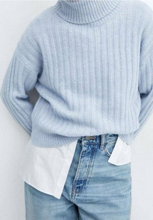 Person iført en lyseblå ribbet rullekrave over en hvid skjorte, kombineret med højtaljede blå jeans mod en ensfarvet baggrund.