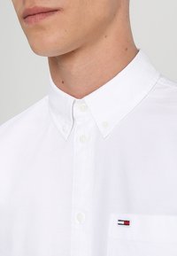 Witte katoenen button-up overhemd met een klassieke kraag, met een klein geborduurd logo in rood, wit en blauw op de borstzak.