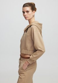 Korte beige hoodie van zachte stof met een rits aan de voorkant, een capuchon met trekkoord, en een geribbelde zoom, gecombineerd met bijpassende beige broek.