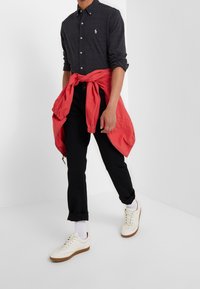 Mörkgrå skjorta med knappar, svarta byxor och vita sneakers med bruna sulor. En röd jacka är knuten runt midjan. Slätt tyg och avslappnad passform.