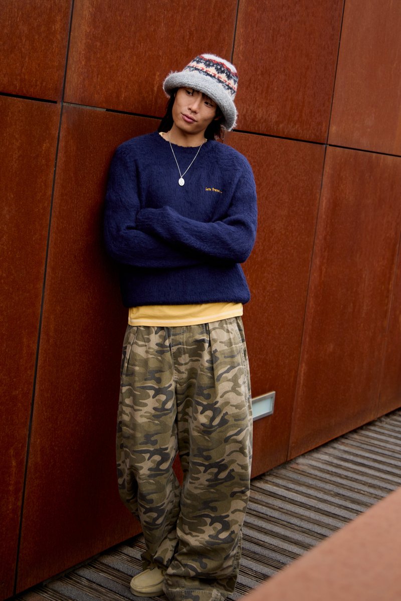 Marineblå fuzzy sweater, gul skjorte og oversized camo bukser. Accentueret med en flerfarvet strikhue og sølv halskæde. Teksturerede overflader synlige.
