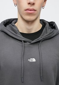 Γκρι φούτερ με κουκούλα και κορδόνι, κατασκευασμένο από μείγμα βαμβακιού. Διαθέτει λευκό κεντητό λογότυπο στο αριστερό στήθος και από κάτω φοράει ένα μαύρο πουκάμισο με στρώσεις.