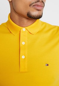 Żółta koszulka polo z teksturowanej dzianiny, z klasycznym kołnierzykiem i trzema białymi guzikami, ozdobiona małym logo na piersi.