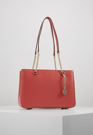 Rode leren DKNY-handtas met gouden kettingbandjes en een ronde gouden merkbedel, tentoongesteld op een wit oppervlak tegen een lichte achtergrond.