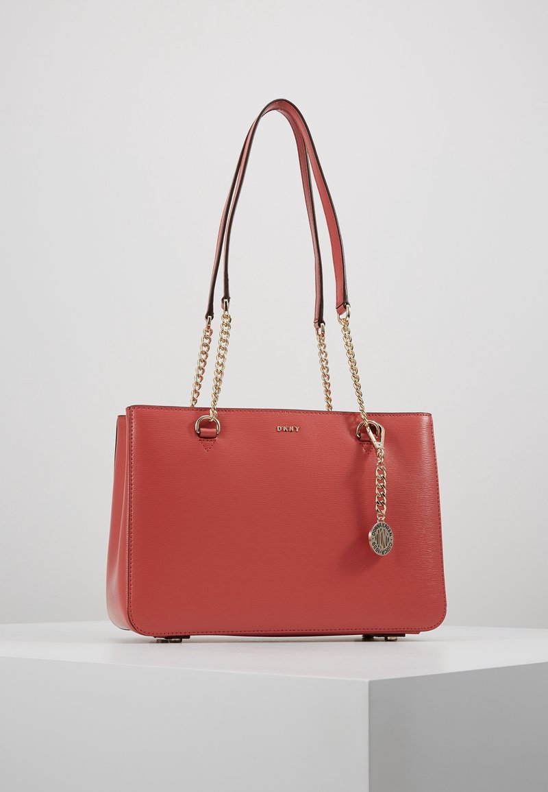 Sac à main DKNY en cuir rouge avec des anses en chaîne dorée et un charm rond doré de la marque, exposé sur une surface blanche sur un fond clair.