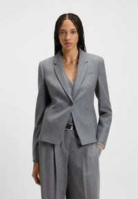 Donna con lunghe trecce che indossa un blazer grigio aderente e pantaloni a vita alta plissettati abbinati, in piedi con una mano in tasca.