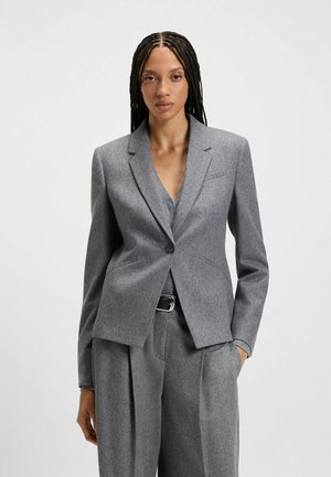 Femme avec de longues tresses portant un blazer gris ajusté et un pantalon plissé taille haute assorti, se tenant avec une main dans la poche.