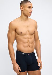 Μυώδης άνδρας με κοντά μαλλιά που φοράει σκούρα μπλε εσώρουχα Tommy Hilfiger, στέκεται μπροστά σε απλό ανοιχτόφωτο φόντο.