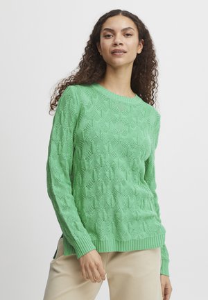 Grön stickad tröja med texturerat diamantmönster, rund halsringning, ribbad mudd och nederkant, bärs över ljusbeige byxor.