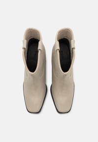 Botines de ante beige con punta afilada, detalle de bordes ondulados y suelas de goma negra. Dos tiradores para facilitar su uso.