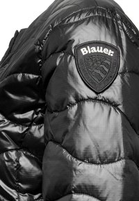 Zwarte gewatteerde jas met een glanzende textuur, voorzien van een kenmerkend schildvormig logo-patch met de tekst "Blauer." Gequilte ontwerp.
