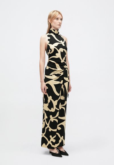 Mulher vestindo um vestido sem mangas com padrões abstratos em preto e bege, gola alta e detalhe assimétrico com laço, combinado com sapatos pretos de bico fino.