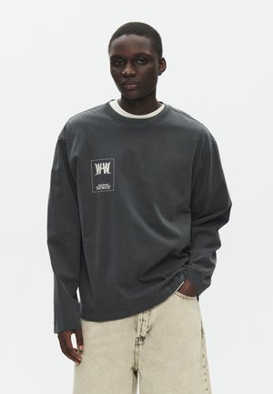 Muškarac u tamnosivoj majici dugih rukava s bijelim logotipom na prsima, u svijetlo bež hlačama, stoji ispred jednobojne bijele pozadine.