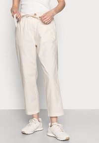 Pantalon beige clair, léger, avec taille élastique, poches latérales et cordon de serrage. Associé à des baskets de couleur claire.