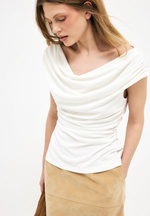 Kobieta z długimi blond włosami ubrana w biały, drapowany, bezrękawnik i beżową spódnicę, patrząca w dół na białym tle.