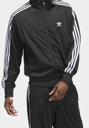 Personne portant une veste et un pantalon de survêtement Adidas noirs avec des bandes blanches sur les manches et les côtés, debout, les bras détendus.