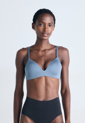 Donna con i capelli corti e ricci, che indossa un reggiseno blu e delle mutande nere a vita alta, in piedi davanti a uno sfondo chiaro e semplice.