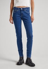 Jeans ajustados de mezclilla azul con una cinturilla de altura media, diseño de cinco bolsillos y detalles de costura sutiles, llevados con botines negros hasta el tobillo.