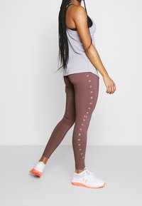 Strój sportowy składający się z szarego topu na ramiączkach i brązowych legginsów z białym napisem logo. Modelka ma na sobie białe buty z pomarańczowymi akcentami. Gładka tekstura tkaniny.