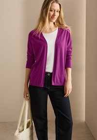 Gilet violet, tricot léger, design à ouverture devant, manches longues, associé à un haut blanc et un jean foncé, tenant un sac de couleur crème.