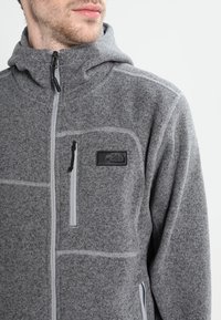 Szary polarowy kurtka z kapturem, pełnym zamkiem z przodu i przeszyciami. Zawiera kieszeń na piersi z czarną łatą logo.