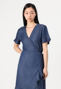 Robe portefeuille en denim bleu profond avec des manches courtes à volants, un décolleté en V et une ourlet asymétrique fluide. Texture lisse avec des coutures subtiles.