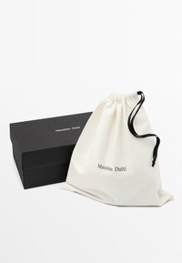 Scatola nera rettangolare di scarpe Massimo Dutti accanto a un sacchetto in tessuto bianco con coulisse nera, entrambi con il logo "Massimo Dutti".