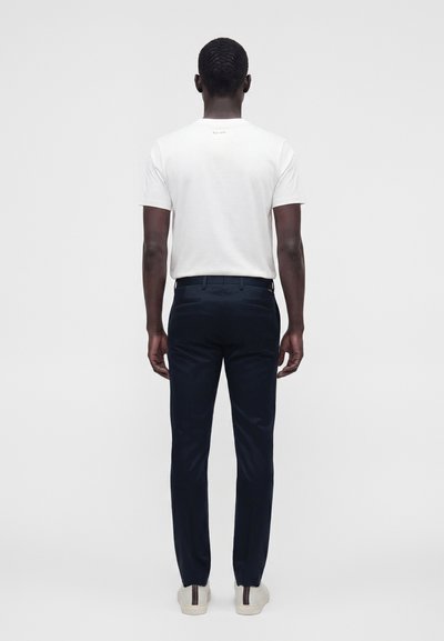 Homme de dos portant un t-shirt blanc uni à manches courtes, un pantalon bleu marine foncé et des baskets blanches avec une bande foncée.