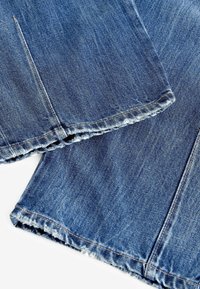 Jean en denim bleu avec ourlets effilochés et coutures visibles, posé à plat avec les jambes de pantalon qui se chevauchent sur un fond blanc.