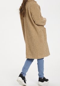 Abrigo beige, esponjoso y sobredimensionado con una superficie texturizada, bolsillos laterales y collar, combinado con jeans azul claro y zapatillas negras.