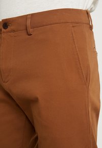 Bruine katoenen broek met een gladde textuur, voorzien van een zijzak, ceintuurlussen en een enkele knoopsluiting.