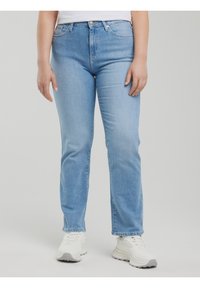 Inimene, kes kannab helesiniseid sirge lõikega teksaseid ja valgeid tossusid, seisab ühevärvilise heleda halli tausta ees.