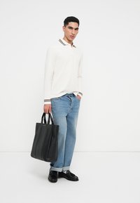 Neste look, uma mala tote preta em couro com costuras verticais é combinada com uma camisa polo de manga longa de cor clara e jeans de denim azul. Calçado casual completa o conjunto.