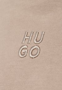 Beige stof met een fijne ribbeltextuur, voorzien van een geborduurd "HUGO" logo in lichtbeige. Het ontwerp is minimalistisch zonder extra accenten.