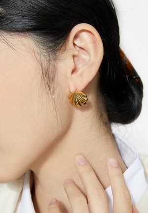 Femme portant une boucle d'oreille clou en forme de coquillage doré, touchant son cou, avec les cheveux attachés en arrière à l'aide d'une pince écaille.