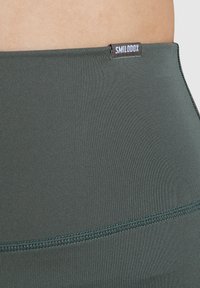 Legginsy o wysokim stanie w ciemnozielonym kolorze, wykonane z gładkiego, elastycznego materiału o minimalistycznym designie; posiadają małą metkę marki przy pasku.