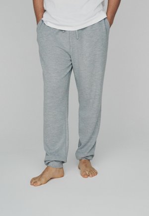 Persona valkā gaiši pelēkas, brīvi pieguļošas bikses un baltu krekls, stāv basām kājām ar rokām kabatās uz vienkrāsaina fona.