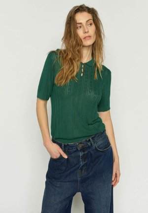 Mujer con cabello largo y ondulado, vestida con un polo verde de manga corta de punto y jeans sueltos azul oscuro, posando con una mano en el bolsillo.