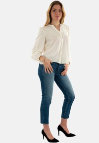 Blusa blanca de manga larga con cierre de botones y detalles plegados, combinada con jeans ajustados azules y tacones de aguja negros.